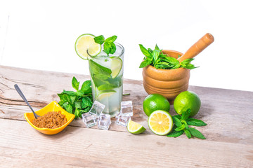 Mojito cocktail with ingredients. Fresh lime, mint leaves and ice cubes, morter with mint and brown sugar on a wooden background. Refreshing alcohol drink. Isolated on white background..