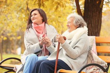 Senior woman with cane and young caregiver sitting on bench in park