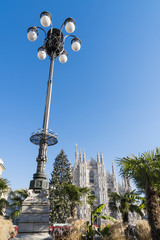 Fototapeta na wymiar Milan, Italy: street lamp in Duomo square with palm garden, Christmas tree and the cathedral facade in a sunny day of december.