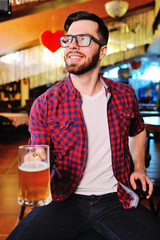 Cute young man in a plaid shirt drinking beer sitting on a chair in a pub