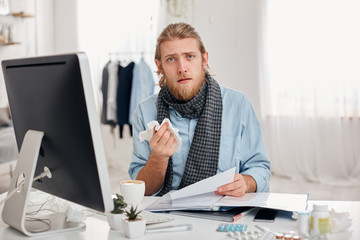 Fototapeta na wymiar Ill bearded man sneezes, uses handkerchief, feels unwell, has flu. Sick male office worker has fever and tired expression, discusses working issues with colleagues. Illness and infection concept