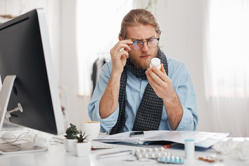 Bearded sick male office worker with spectacles on reads prescription of medicine. Young manager has bad cold, sits at table with pills, tablets, vitamins and drugs on its surface. Health problems