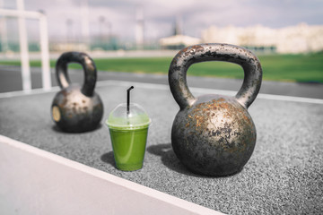 Naklejka na ściany i meble Health and fitness green smoothie detox drink at gym with kettlebells weights at outdoor training fitness center. Plastic cup of vegetable juice morning breakfast next to kettlebell weight equipment.