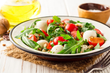 Fresh salad with arugula, cherry tomatoes, mozzarella cheese and walnuts on white wooden background.
