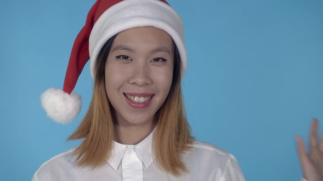 close up portrait young asian woman waving hand say hello wearing santa claus hat on blue background in studio. attractive millennial looking at the camera