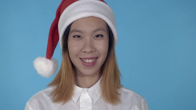 close up portrait young asian woman shows symbol okey wearing santa claus hat on blue background in studio. attractive millennial looking at the camera