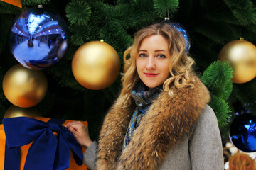 Young beautiful girl in a winter coat on the background of the Christmas tree decorated with balls in a shopping center close-up