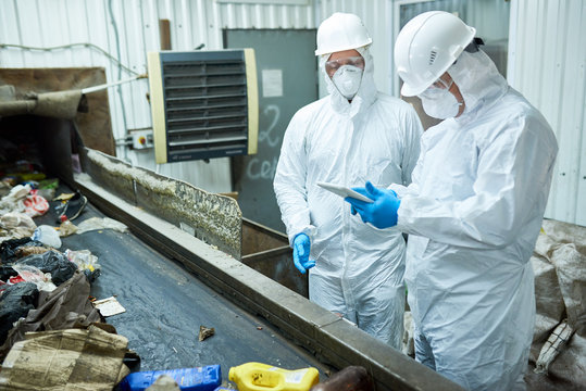 Portrait of two workers  wearing biohazard suits working at waste processing plant sorting trash and using digital tablet standing by conveyor belt, copy space