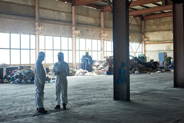 Full length portrait of two workers wearing warehouse suits standing in empty workshop of waste processing plant, piles of trash in background, copy space