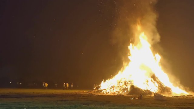 Big bonfire burning in the field at night, people wearing safety vests and helmets standing next to it