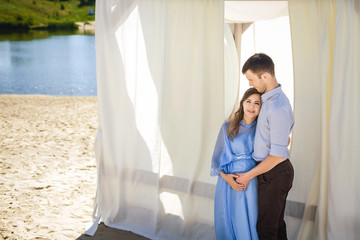 Obraz premium future mother with curly long hair in beautiful blue dress and handsome father hugging near light alcove on beach, happy family concept