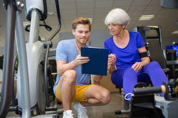 trainer with clipboard helping eldery woman in gym