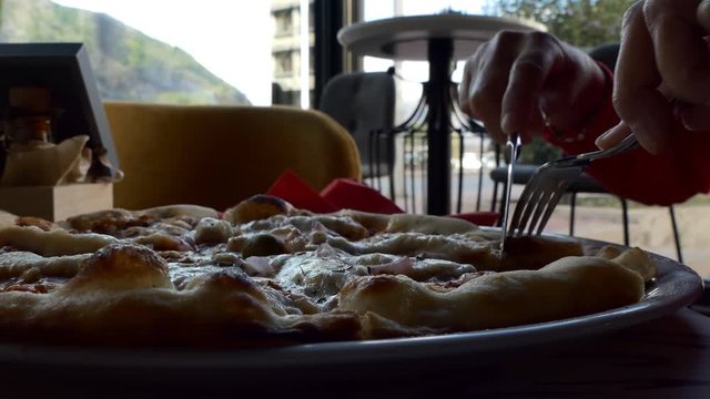 Close up of plate with pizza being cut by woman with knife and fork on table in cafe by the street in town.Selective focus on the meal