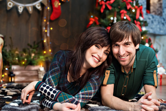 Beautiful couple in green on a background of Christmas decorations. Happy family in a New Year's loft with bokeh lies on the carpet, hugs, behind the back is a fireplace and a tree. Copy space card