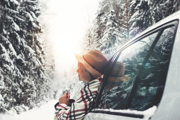 Beautiful woman in vintage hat and checkered scarf enjoying a winter view of the road from the car window. Taveler by car rides on a snowy road