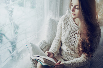 Young beautiful girl wearing knitted dress sitting on windowsill, holding and reading book. Day...