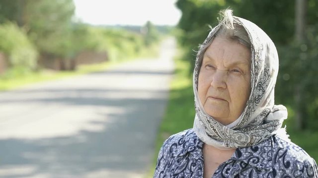 Thoughtful look of serious mature elderly woman aged 80s with gray hair dressed in a handkerchief on the background of road in summer. Slow Motion