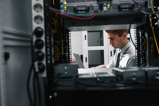 Portrait of modern young man holding laptop standing in server room working with supercomputer