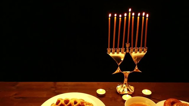 on the table is a Hanukkah candlestick with burning candles. on the table there are donuts, cakes, honey and chips. the camera moves from left to right.