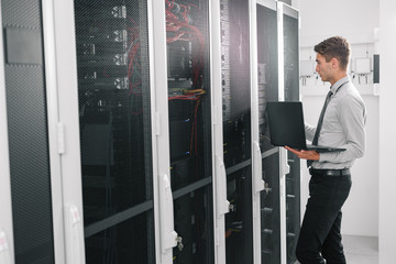 Portrait of modern young man holding laptop standing in server room working with supercomputer