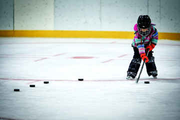 A kindergarten aged little girl in ski pants and winter jacket and helmet lining up a row of hockey pucks to practice shooting at a net