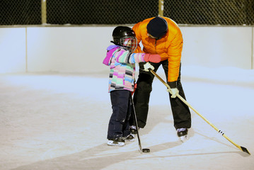 An adult male with beard helping a little girl to learn to skate on an outdoor ice skating rink