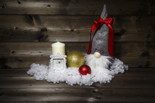Christmas decoration with white candle, picture frame, shiny Christmas balls, snow and scandinavian traditional gnome tomte, nisse, tomtenisse, tonttu with a red ribbon on a wooden background