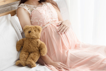 Pregnancy woman  in pink dress and teddy bear  on the bed in bedroom.