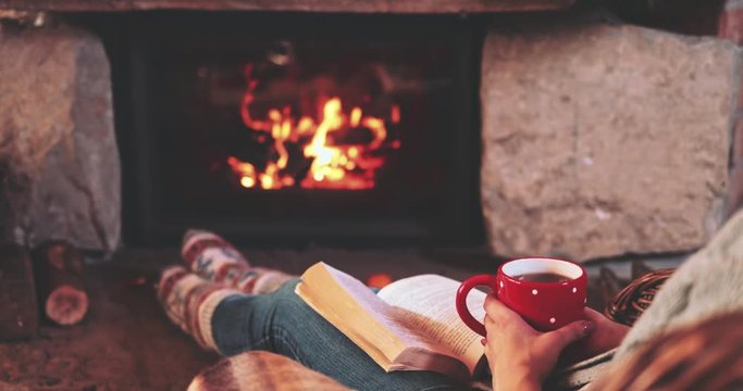 Woman relaxes by warm fire with a cup of hot drink and warming up her feet in woollen socks. Feet in woollen socks by the Christmas fireplace. Cozy atmosphere. Winter and Christmas holidays concept.