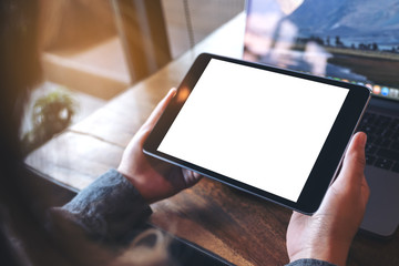 Mockup image of hands holding black tablet pc with white blank screen and laptop on wooden table background in cafe