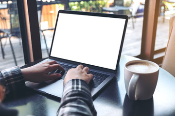 Mockup image of business woman using and typing on laptop with blank white screen and coffee cup on wooden table in cafe