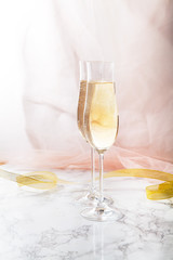 Two flute glasses with champagne on marble background. Pink transparent fabric. Soft light
