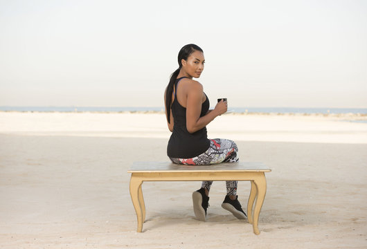 Beautiful Biracial Athletic woman wearing a sports outfit sits by the beach smiling at you drinking coffee or tea enjoying the bright sunny morning sun  - Powered by Adobe
