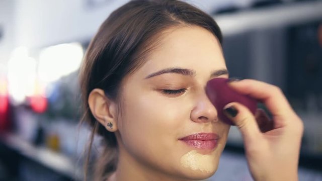 Unrecognizable visagist applying fluid foundation on young woman's face using sponge. Professional makeup in salon. Slowmotion shot
