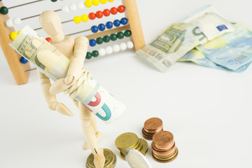 Eine Holzfigur trägt ein Bündel Geldscheine, im Hintergrund ein Rechenschieber, daneben noch einige Geldscheine und Geldmünzen