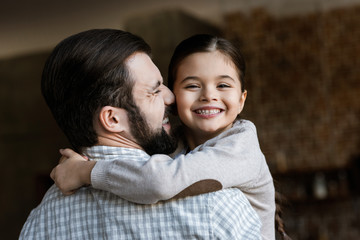 cheerful father and daughter hugging and looking at camera at home