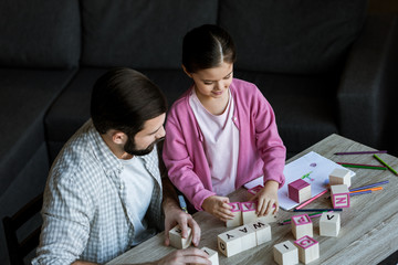 father with daughter sitting at table and making words by cubes with letters at home