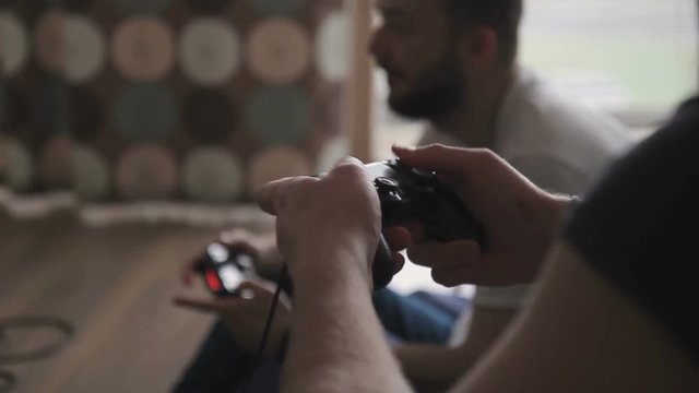 Two men play in game, holding game consoles in room indoors. Young gamer act with enthusiasm, pressing fingers on buttons of black compact device with wire that they have in hands. They sit on sofa in