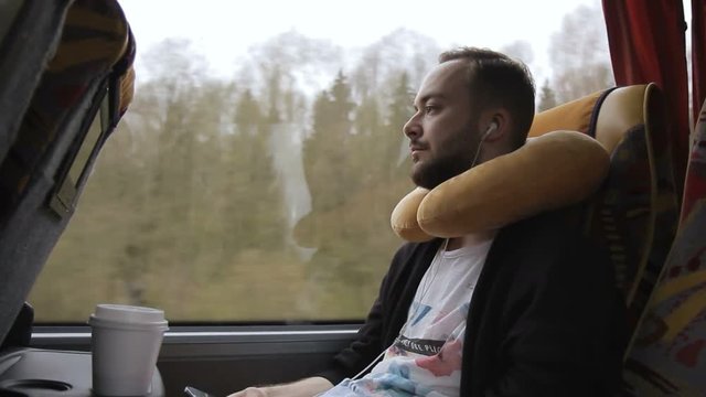 Man with a pillow chooses music for listening during a bus trip. Bearded passenger with headphones presses his fingers on smartphone screen while traveling by window.