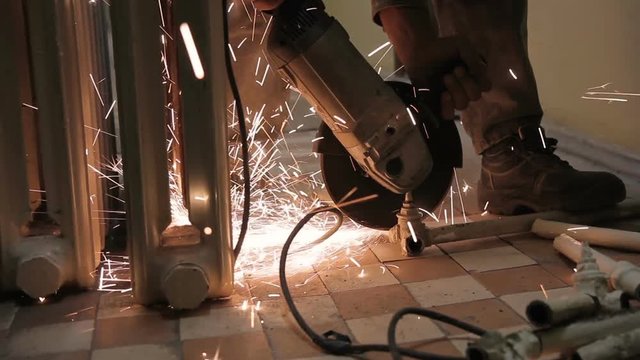 Man holds a saw in his hands and cuts details from old metal battery. Worker with bright flashes and sparks divides the white structure in the room into parts.