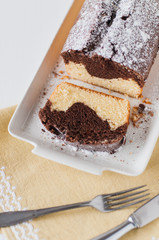 Slice of traditonal German vanilla and chocolate marble cake with whole cake on white cake serving platter with fork and knife on vintage, yelow table cloth