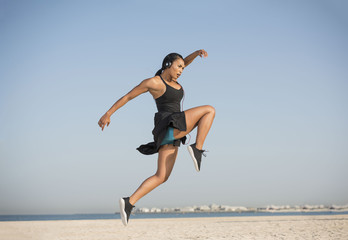 Beautiful Biracial woman by the ocean on a clear Summer morning does an expressive dance move wearing shorts and sports top as she listens to music on her headphones 