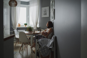 A young woman with a baby sitting in a cozy grey kitchen and looking out the window. Front of her round table and white chairs.