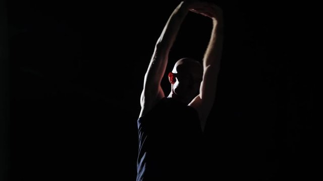 Half-lit silhouette of a man stretching in a darkened room. He is stretching his arms and rotating his upper body with his arms up over his head.