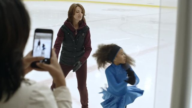 Mother standing at ice rink border and shooting video with smartphone of her little daughter training with figure skating coach