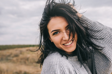 Obraz premium Closeup portrait of happy brunette young woman wearing gray sweater being playful and carefree with beautiful toothy smile and windy hair posing on landscape. Lifestyle concept. Cover idea mood.