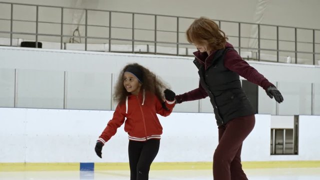 Female coach holding hands with little African girl while skating with her on indoor ice rink