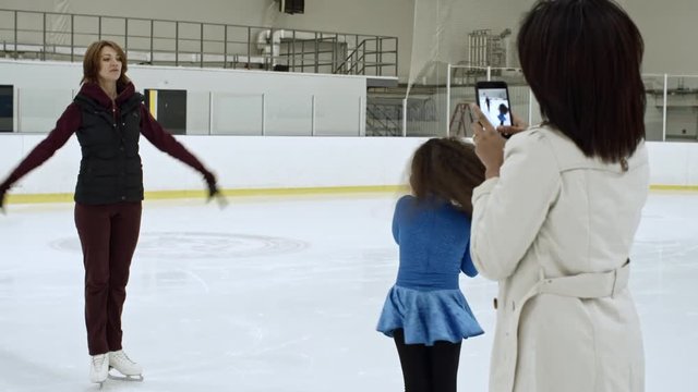 Mother recording video with smartphone of her little daughter spinning on ice in blue dress while training with coach