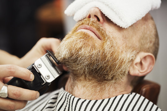 A customer sitting in the barber's chair, with a hot towel on his face, and a barber trimming his beard with an electric trimmer. 