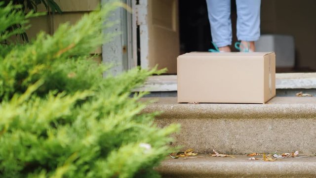 The woman takes the parcel from the threshold of her house. In the frame, only the door, the box and the legs of the woman are visible